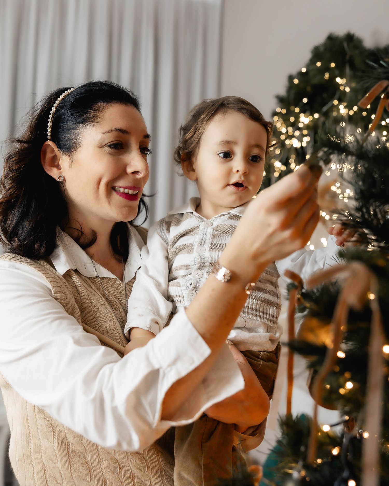 A smiling mother holds her toddler while decorating a Christmas tree with warm fairy lights at home.