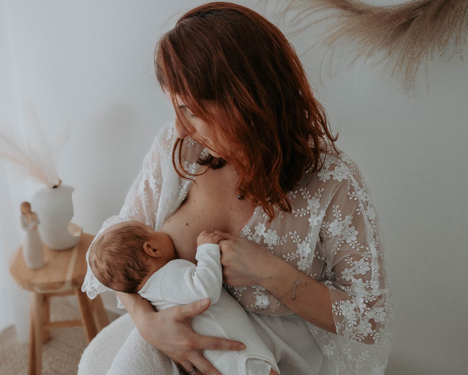A mother with red hair in a lace dress tenderly breastfeeding her newborn baby in a serene, softly lit room.