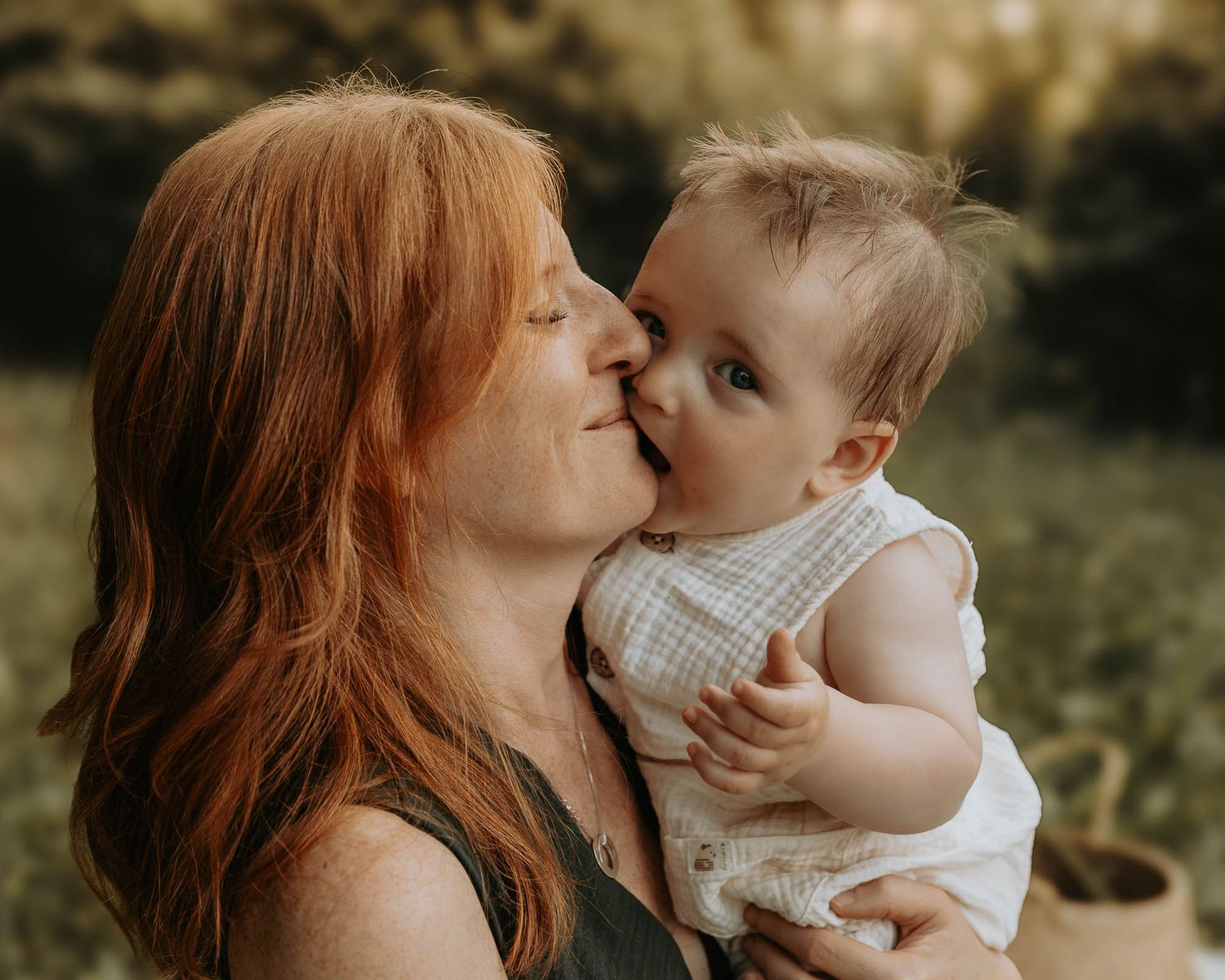 A red-haired mother lovingly kisses her smiling baby outdoors in soft natural light.