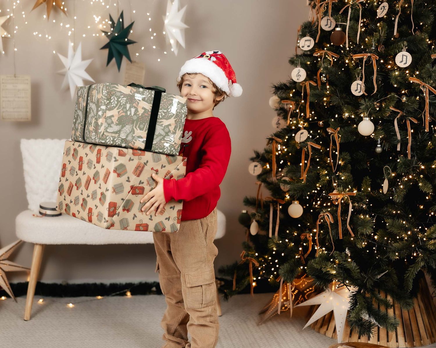 A smiling boy in a Santa hat and red sweater holds wrapped gifts beside a decorated Christmas tree.