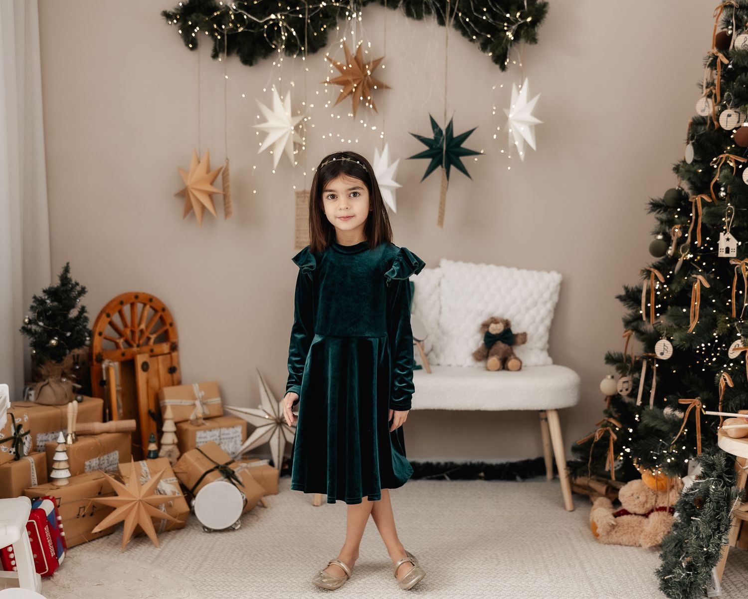 A young girl in a teal velvet dress poses in a festive Christmas room with wrapped gifts and a decorated tree.