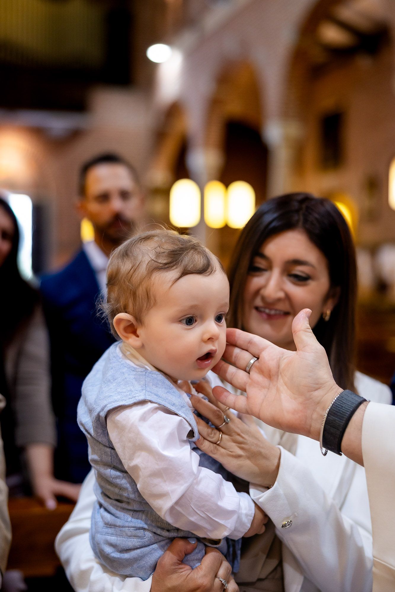 A smiling woman holds a dressed baby during a church baptism ceremony, with guests visible in the background.