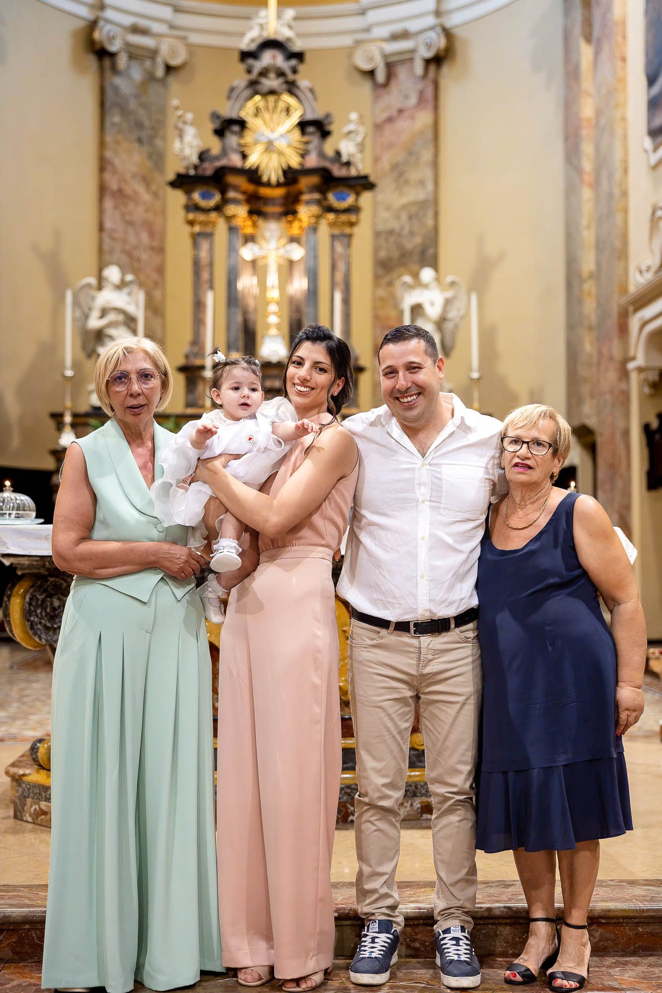 Family posing together in an ornate church during a baptism ceremony, with parents holding their baby.