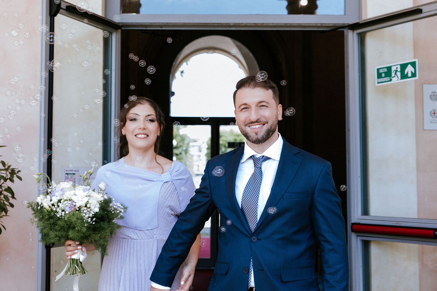 Wedding party in navy suits and light dresses pose together outside a stone building entrance.