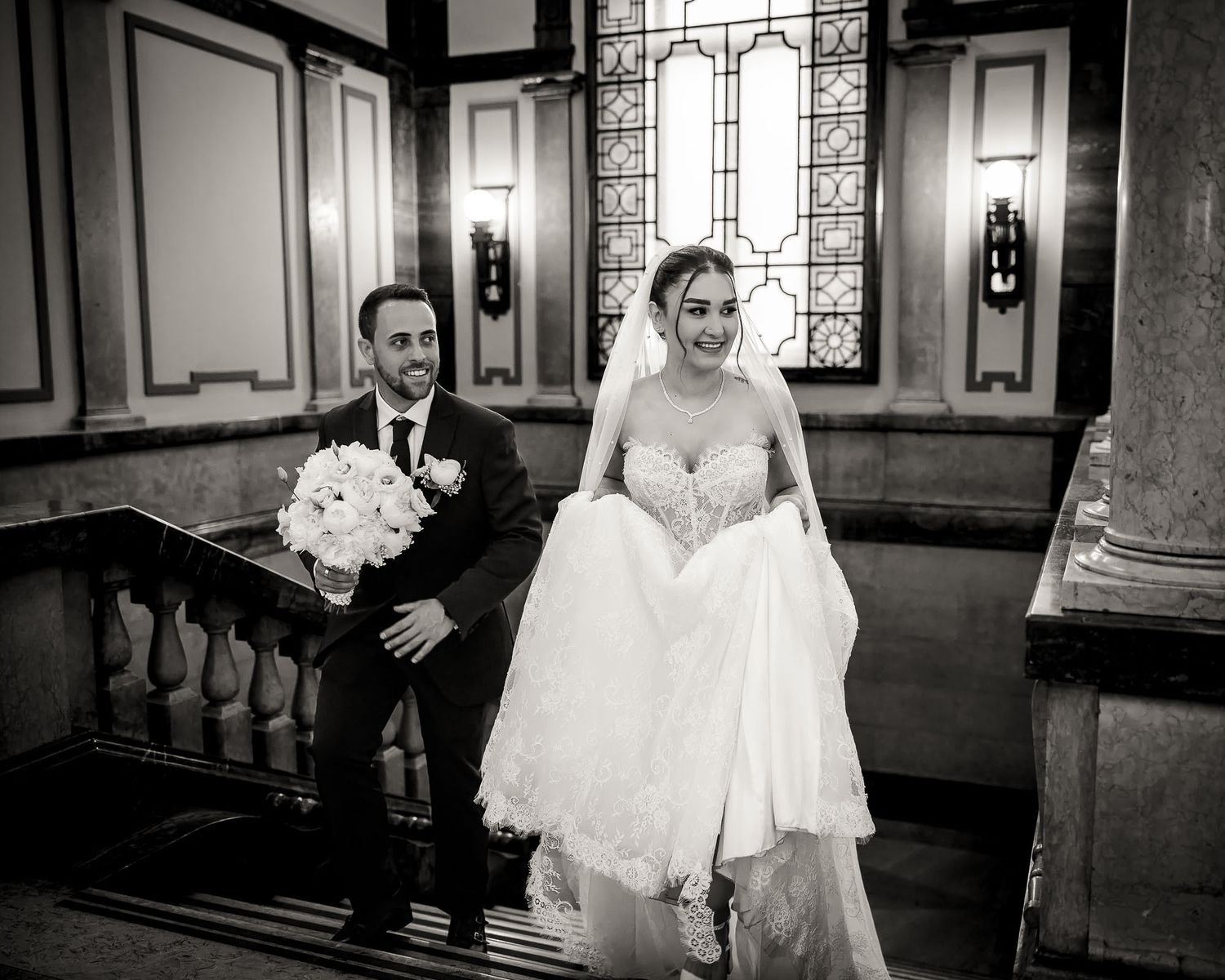 Black and white wedding photo of bride and groom descending grand staircase, bride holding bouquet and dress.