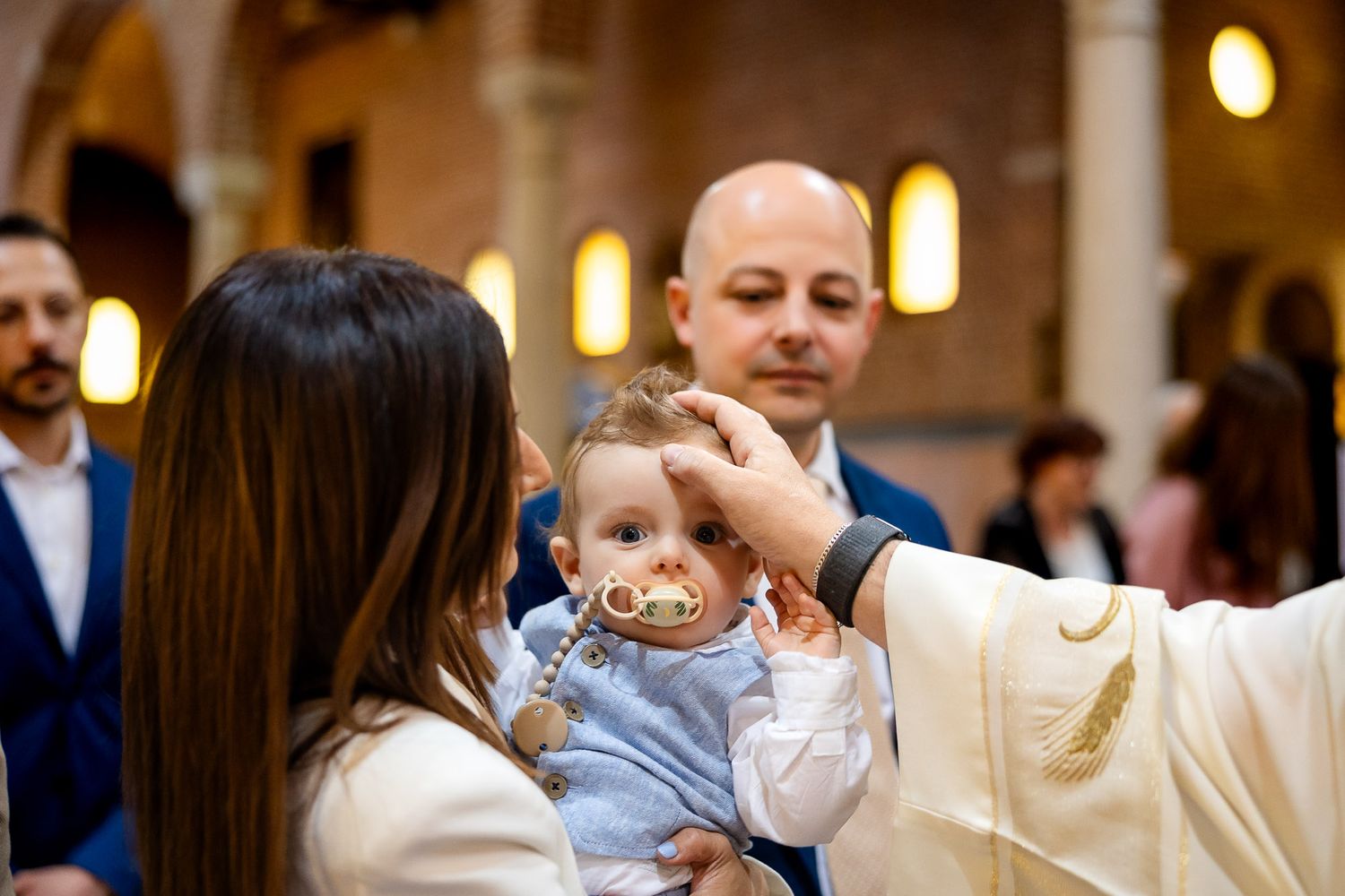 Priest performing baptism on a baby held by mother in a church, with godfather standing behind.