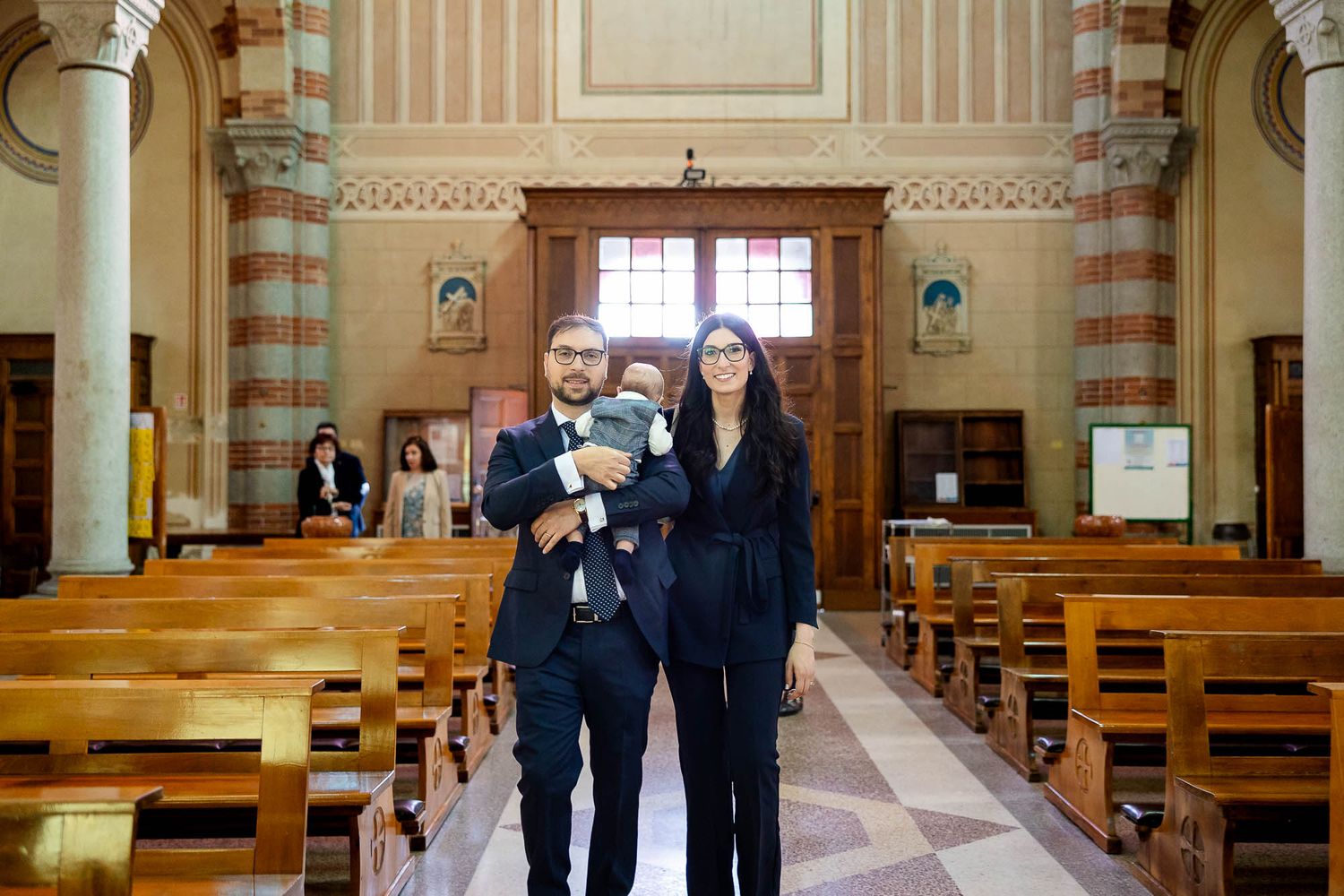 A couple holding a baby stands inside an elegant church with wooden pews and ornate decor.