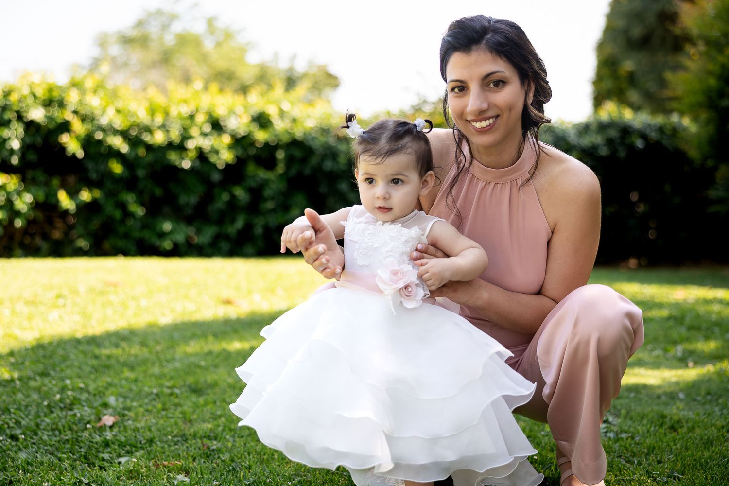 Mother and baby girl in matching pink and white outfits posing together on green garden lawn.