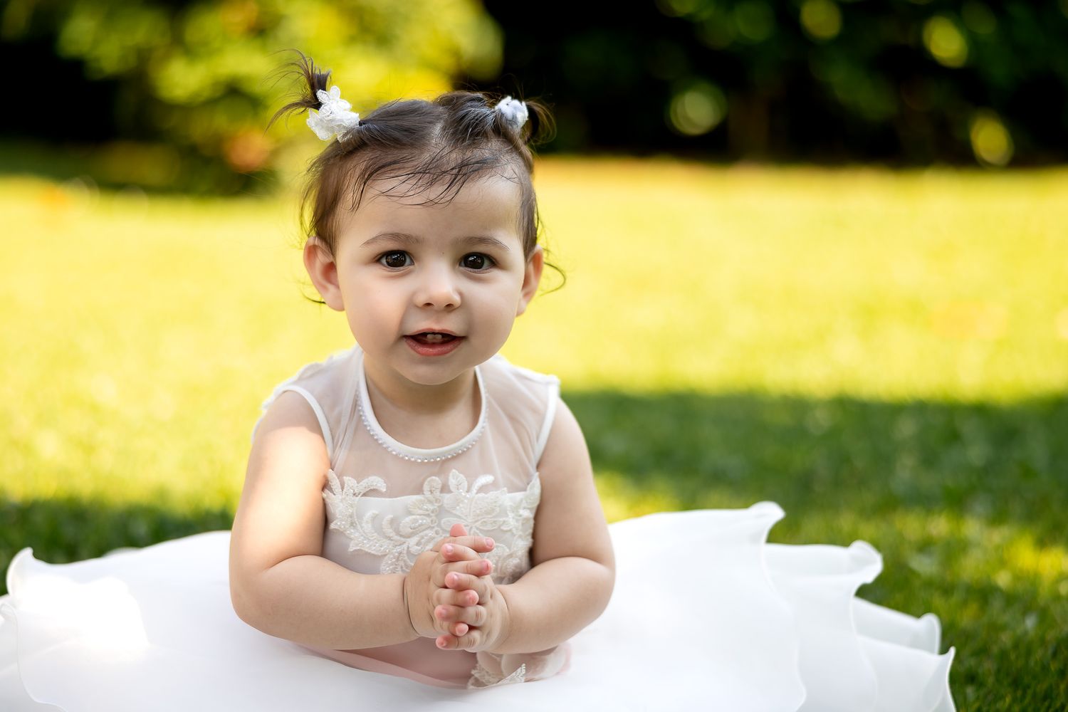 Adorable baby girl in white tutu dress sitting on green grass, smiling with hair accessories outdoors.