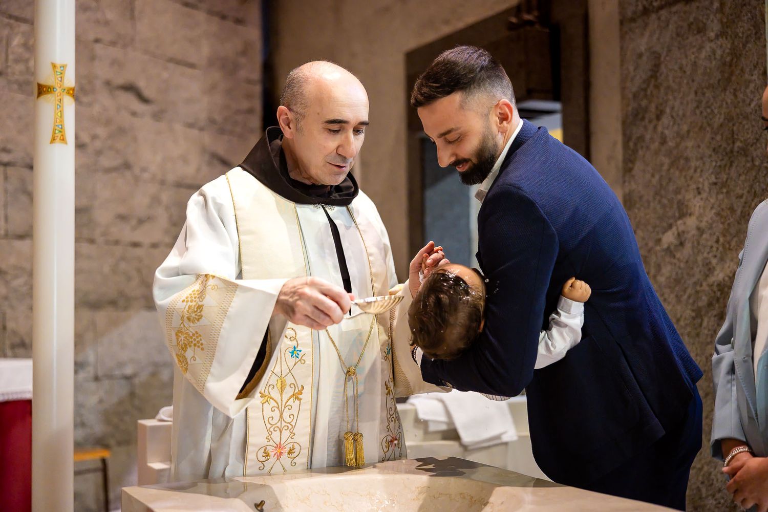 Priest pouring holy water over infant's head during a baptism ceremony at a stone baptismal font.