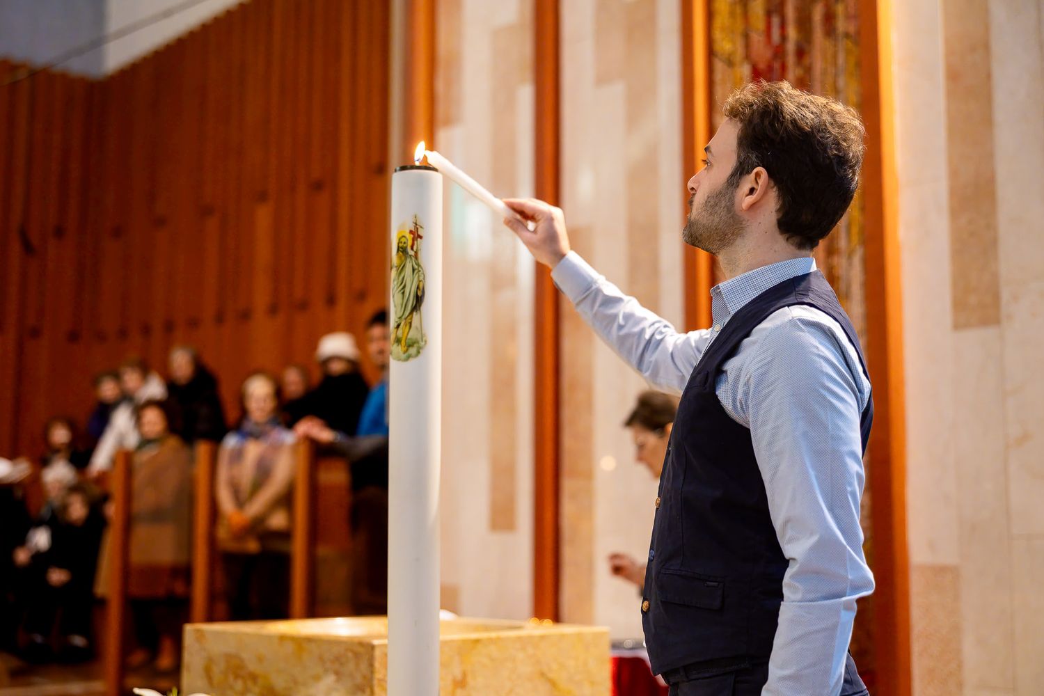 Man lighting a tall white candle on an altar inside a church during a religious ceremony.