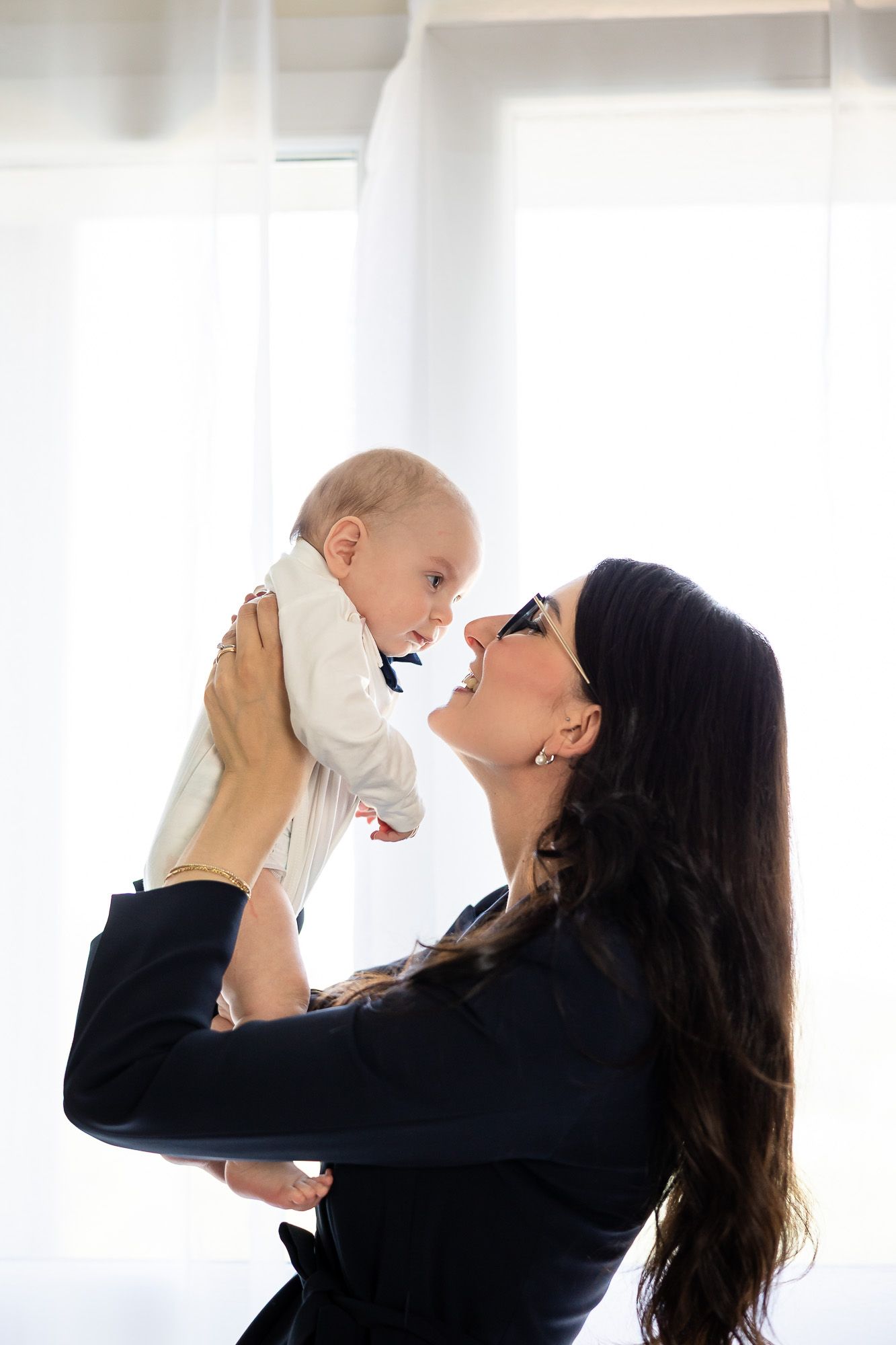 A smiling businesswoman in glasses lifting and bonding with her happy baby in a bright white room.