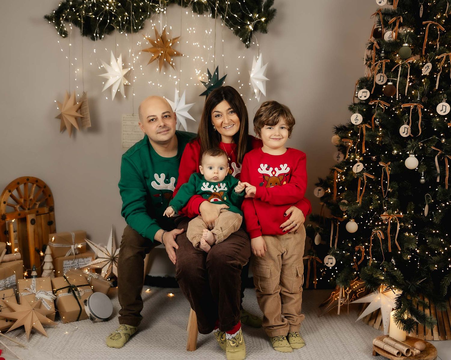 Family of four in matching Christmas sweaters poses together near decorated holiday tree with gifts.