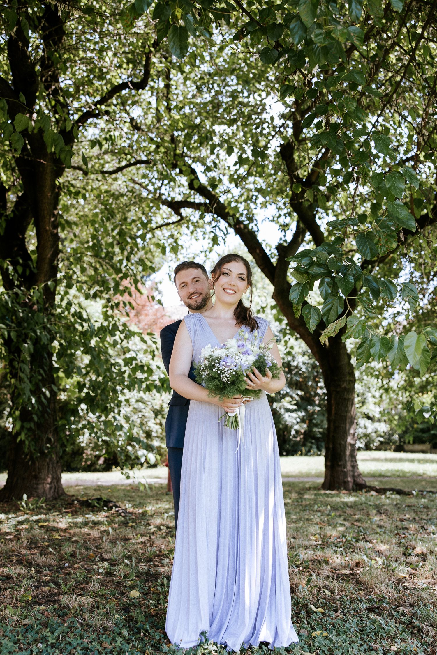 A couple shares a romantic moment outdoors beneath a canopy of green trees in a garden setting.