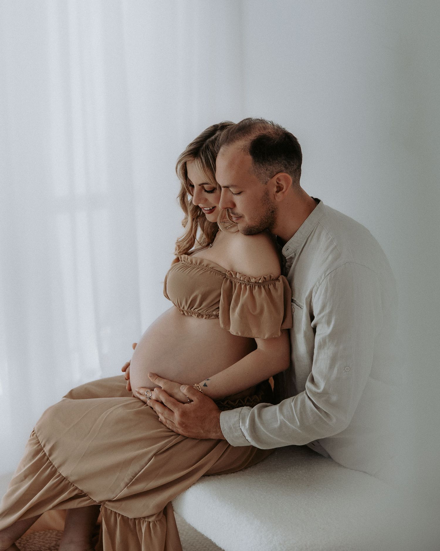 Una coppia condivide un tenero servizio fotografico di maternità: la donna indossa un abito color beige mentre il partner le abbraccia