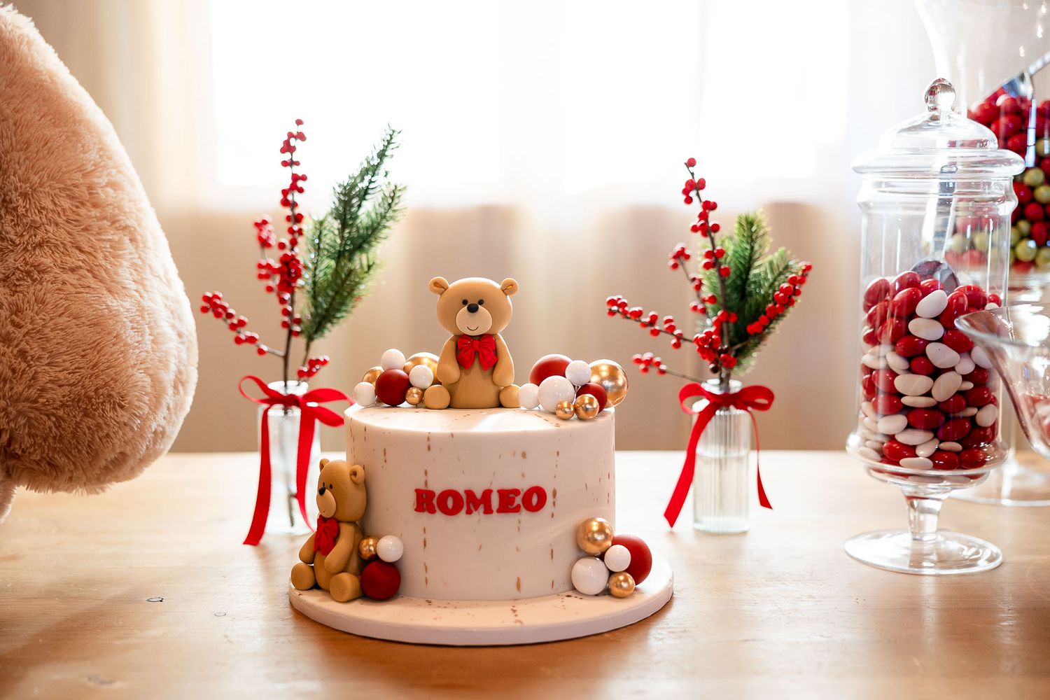 White birthday cake with teddy bear decorations spelling ROMEO, surrounded by red berry branches and candy jars.