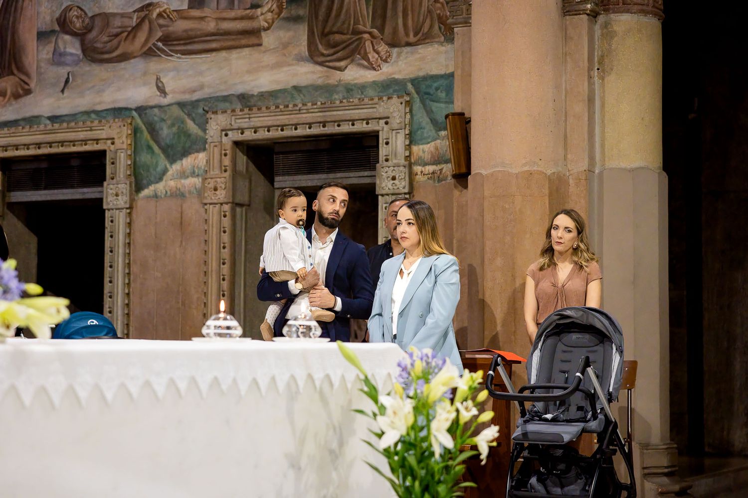 Family gathered at church altar during baptism ceremony, with man holding child, women standing nearby and stroller visible.