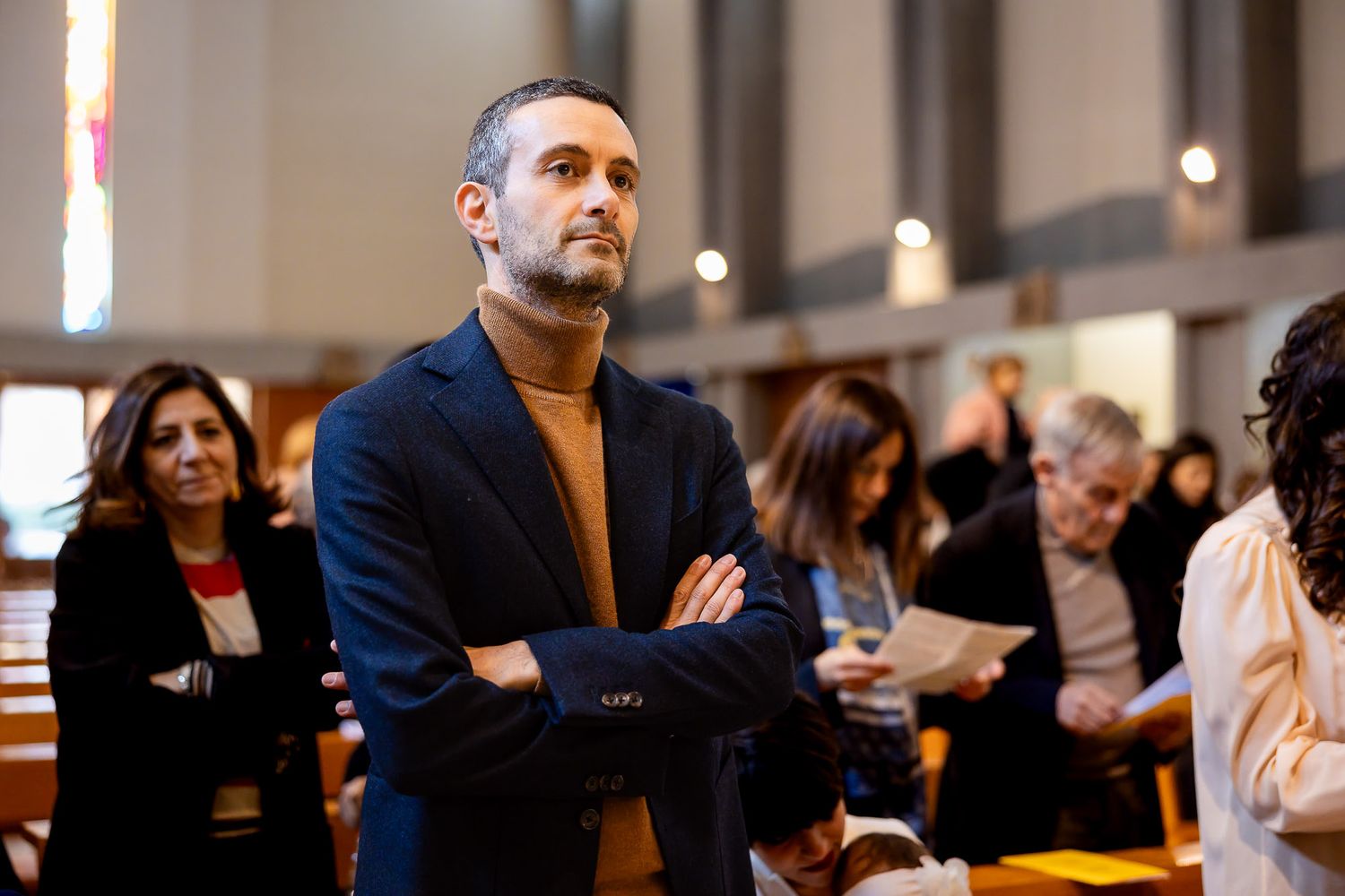 Man with arms crossed standing in a church during a religious service with congregation in background.