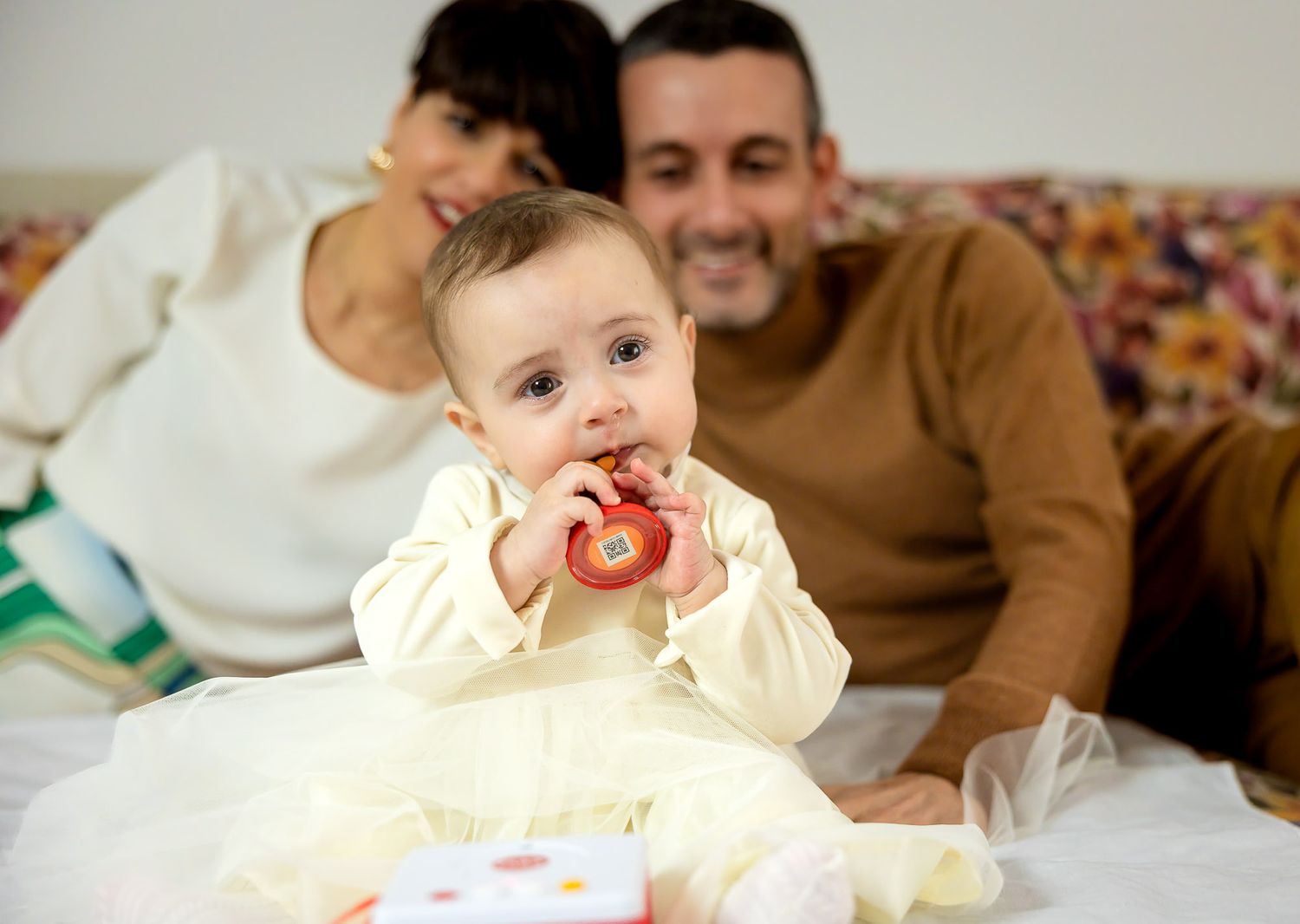A happy baby chews on a colorful toy while smiling parents relax together on a white bed behind them.