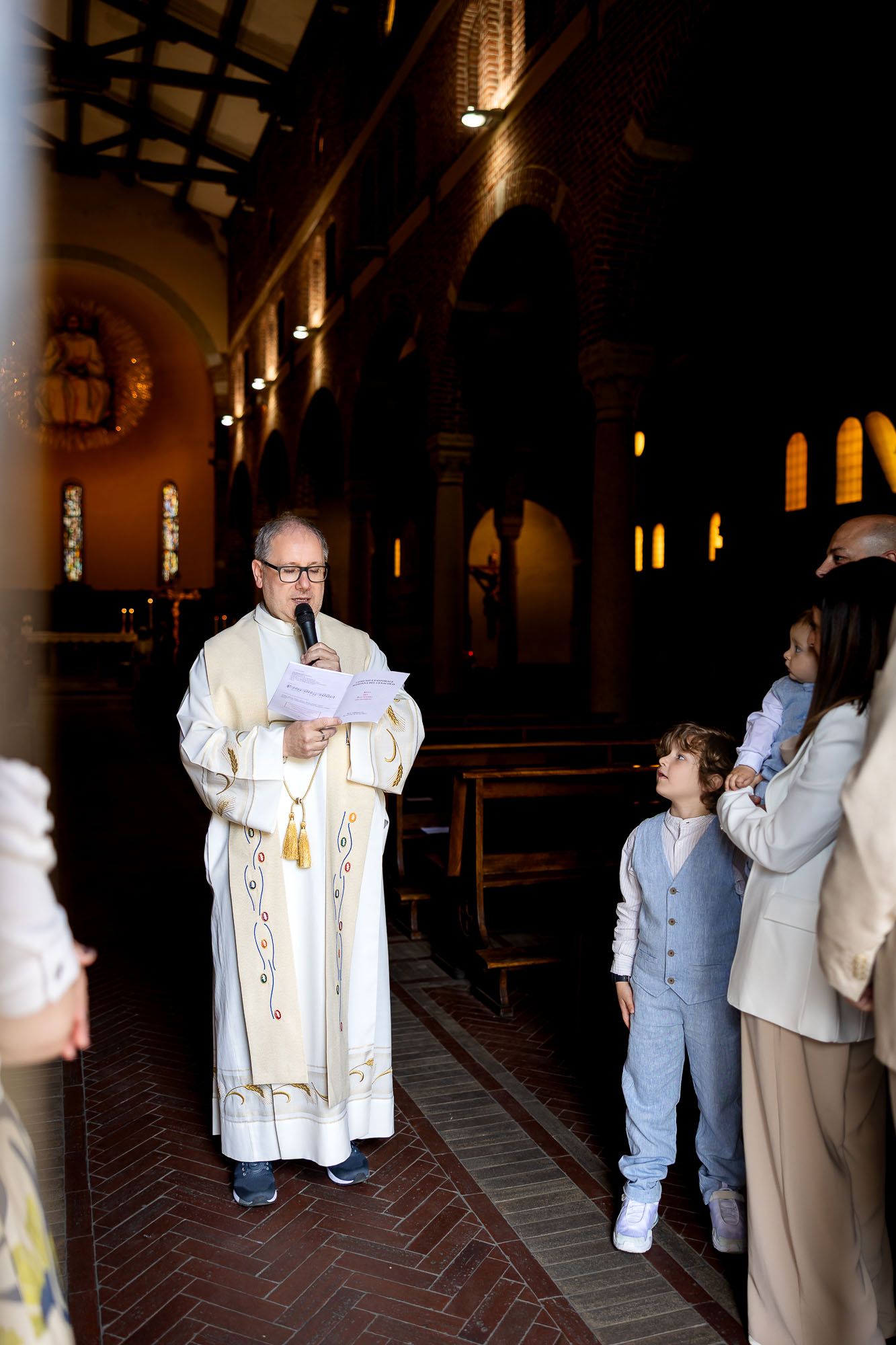 Priest in white vestments reads from papers during a church ceremony while children and family members stand nearby.