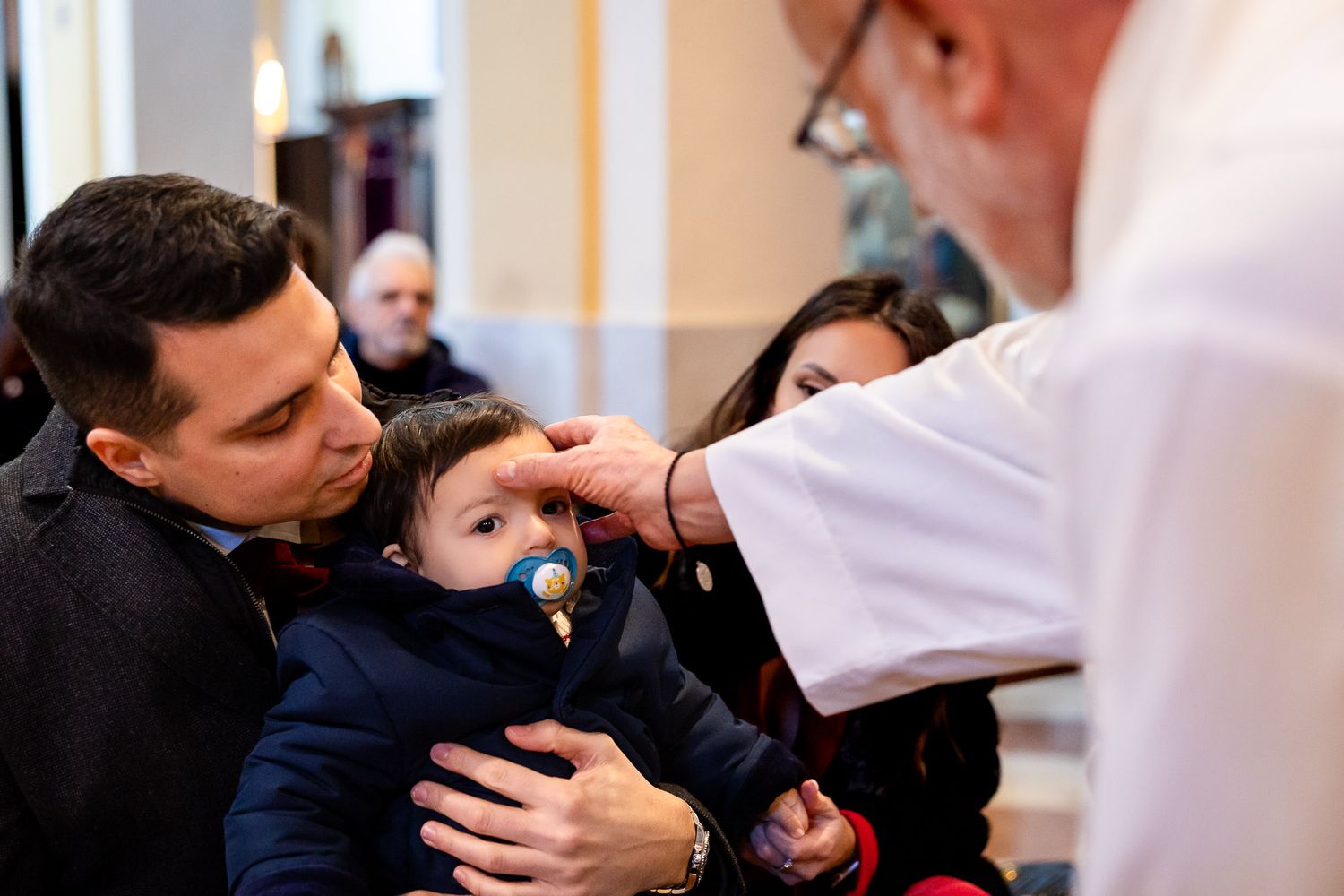 Priest performing baptism blessing on infant with pacifier held by father in church.