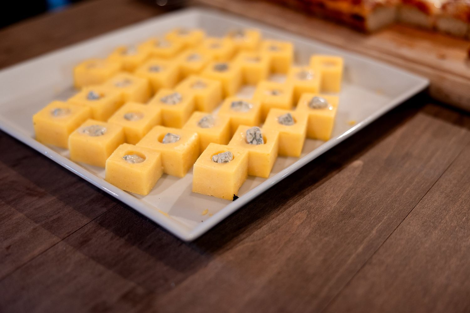 Yellow cheese cubes with herb toppings arranged in rows on a white rectangular serving platter on a wooden board.