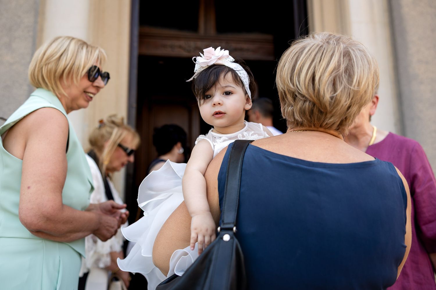A baby girl in a white dress and headband being held by a woman at a church event.