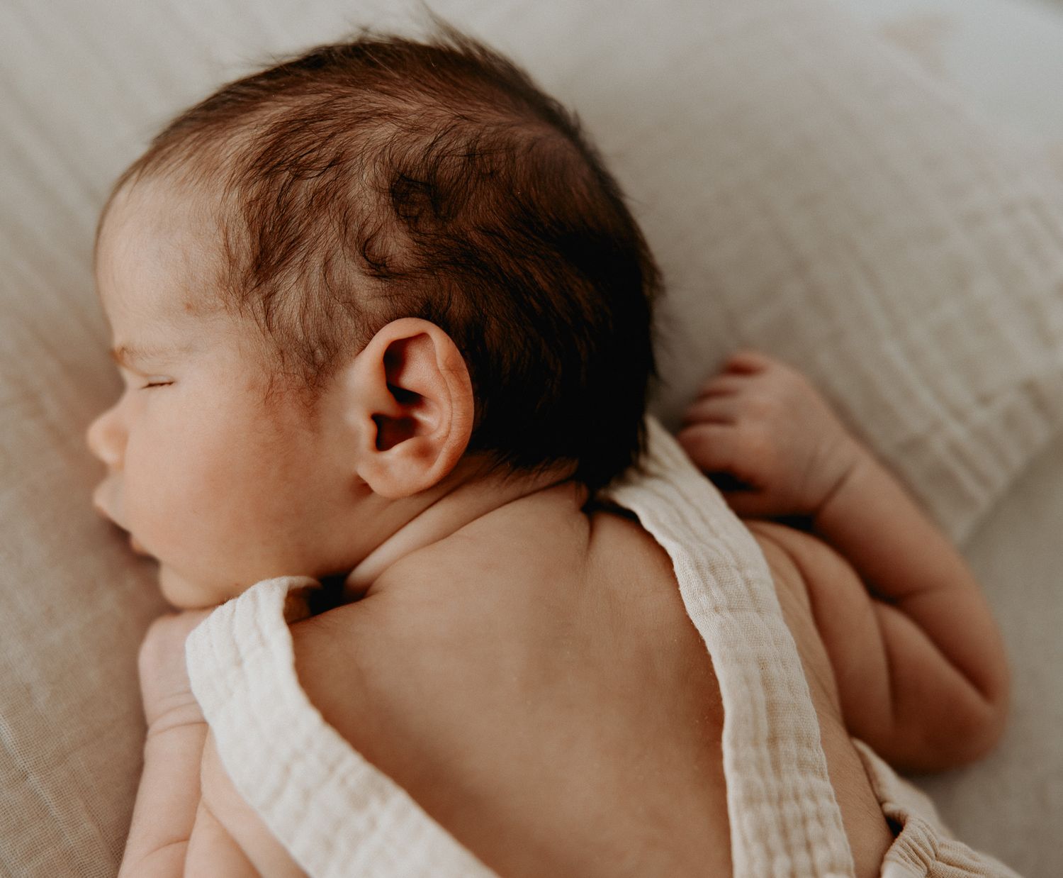 A sleeping baby in a white onesie rests peacefully on soft bedding, viewed from behind.