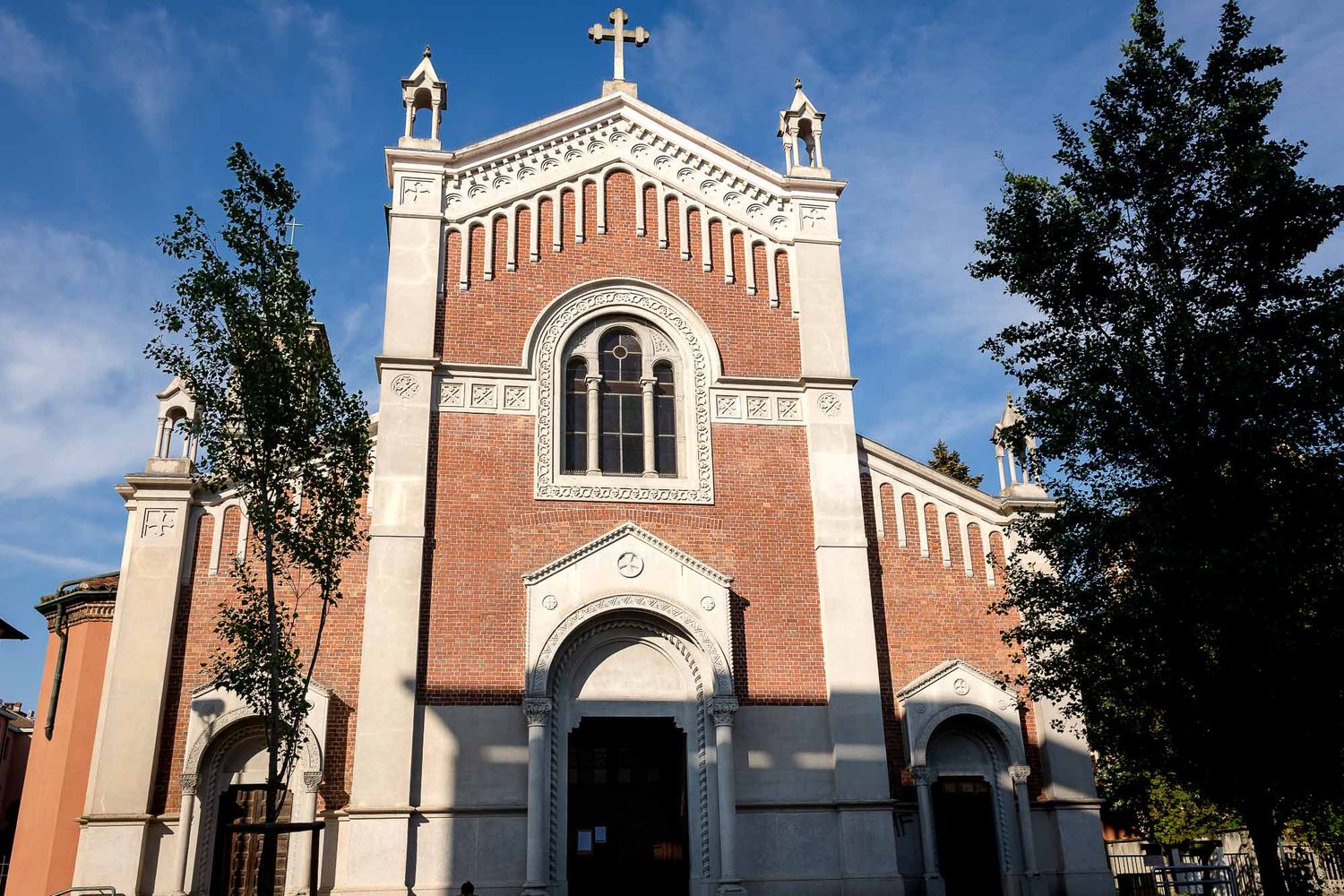 Historic red brick Catholic church facade with arched windows, cross on top, and trees under blue sky.