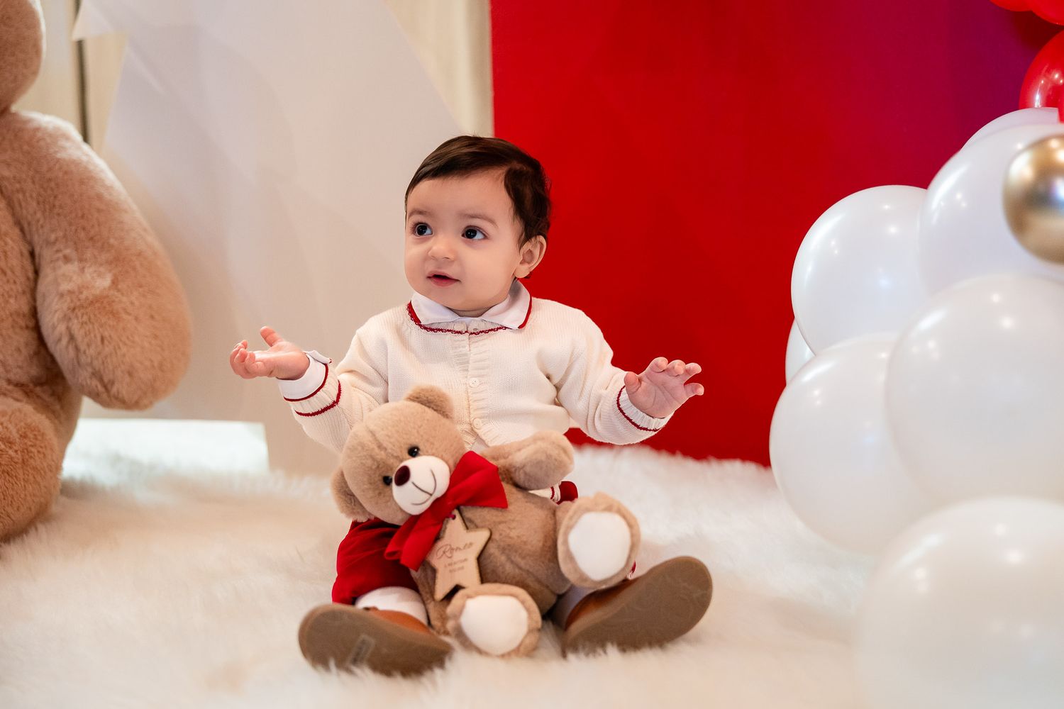 Adorable baby in white outfit sits with teddy bear on fluffy rug, red backdrop with white balloons.