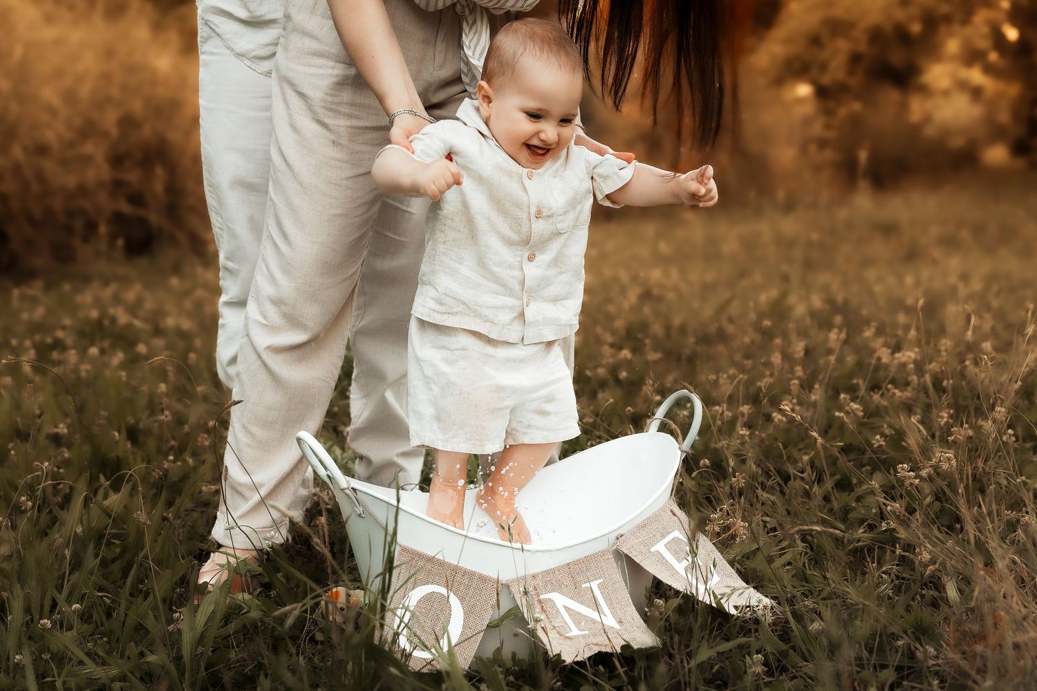 A baby stands in a white bucket outdoors, supported by a parent, with the word ONE overlaid on the golden field background.