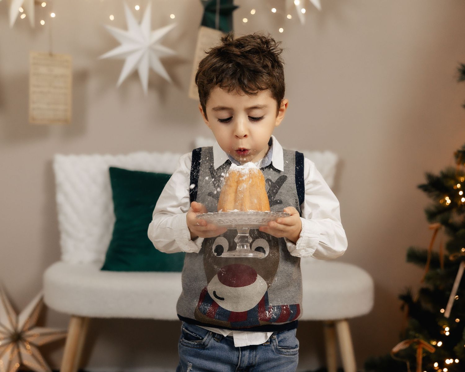 A young boy in a festive vest blows on a bundt cake held in a reindeer box amid Christmas decorations.