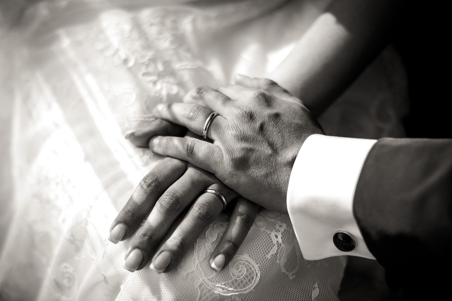 Black and white photo of bride and groom hands with wedding ring and cufflink on lace dress.