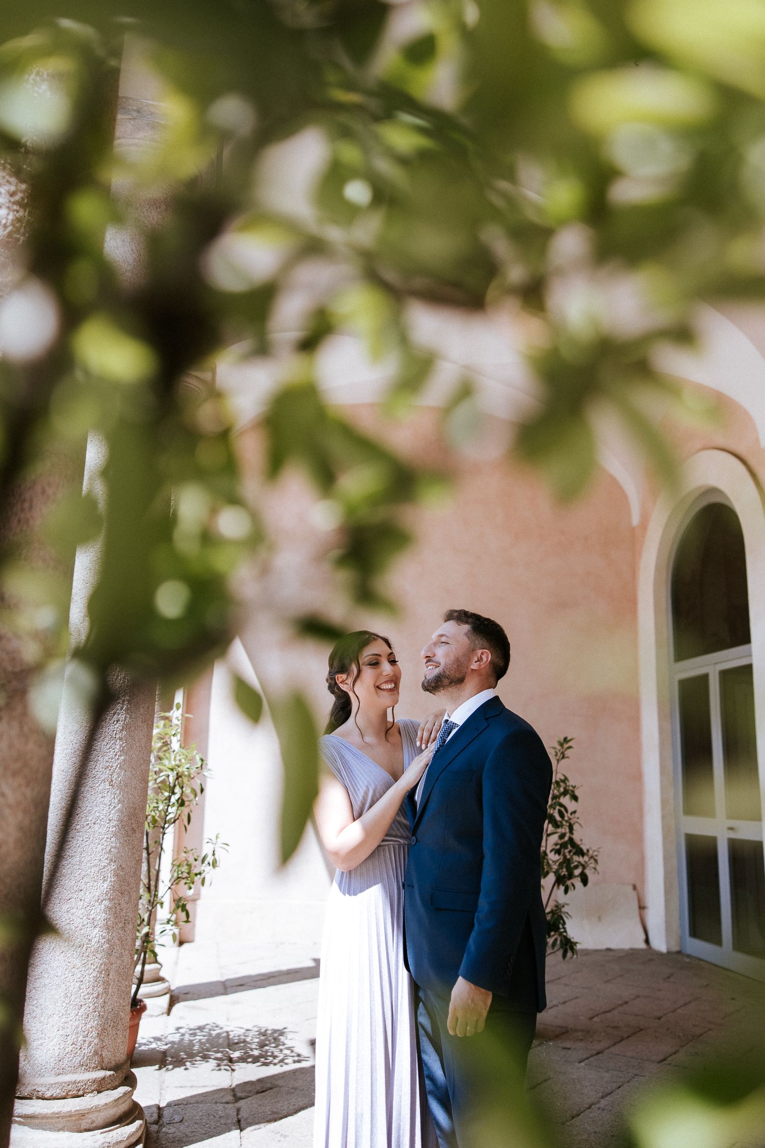 A couple shares a romantic moment in an elegant outdoor corridor with arched windows and soft natural lighting.
