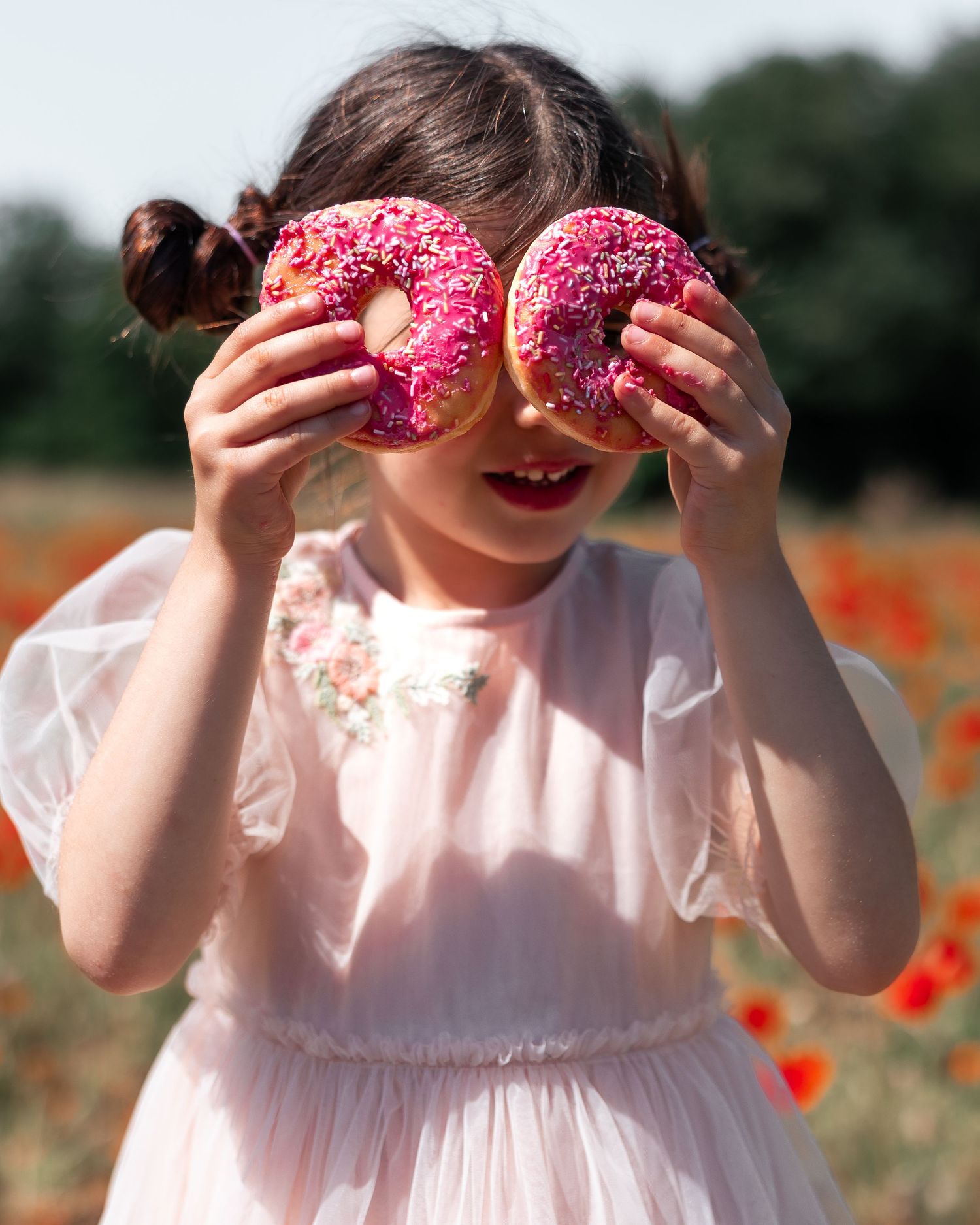 Child in white dress holding pink donuts over eyes in blooming poppy field.