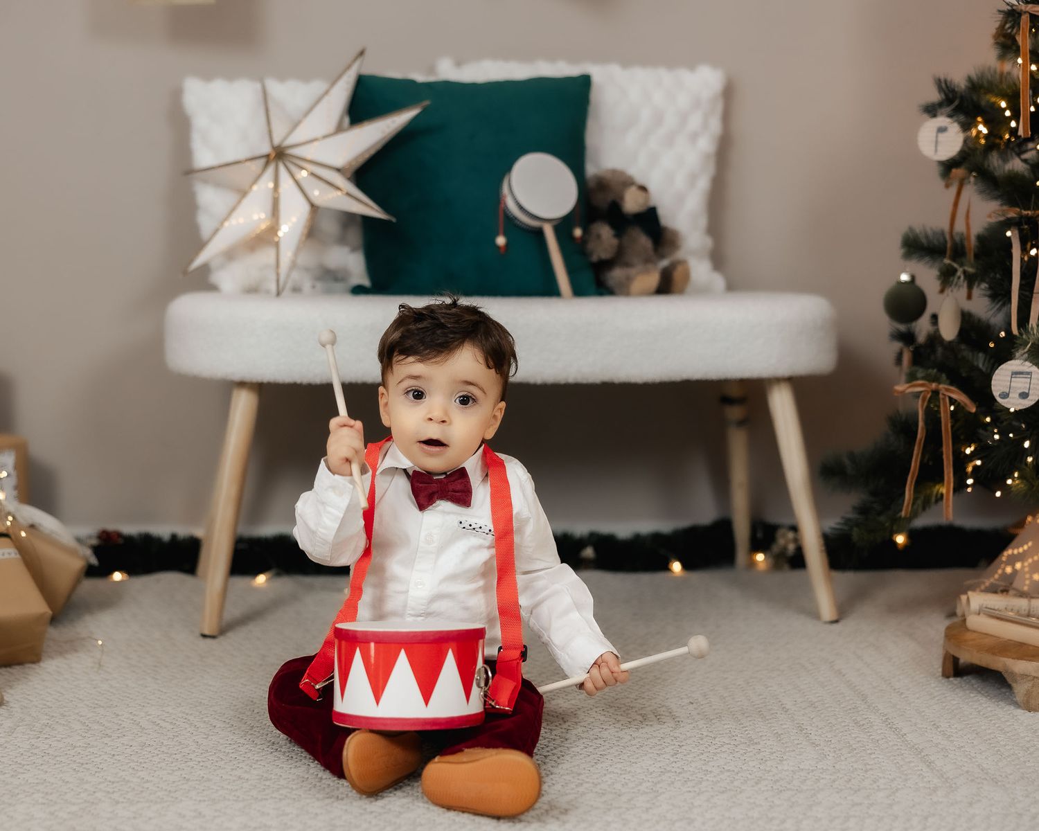 Adorable baby boy in suspenders and bow tie plays with toy drum near Christmas tree holiday decor.
