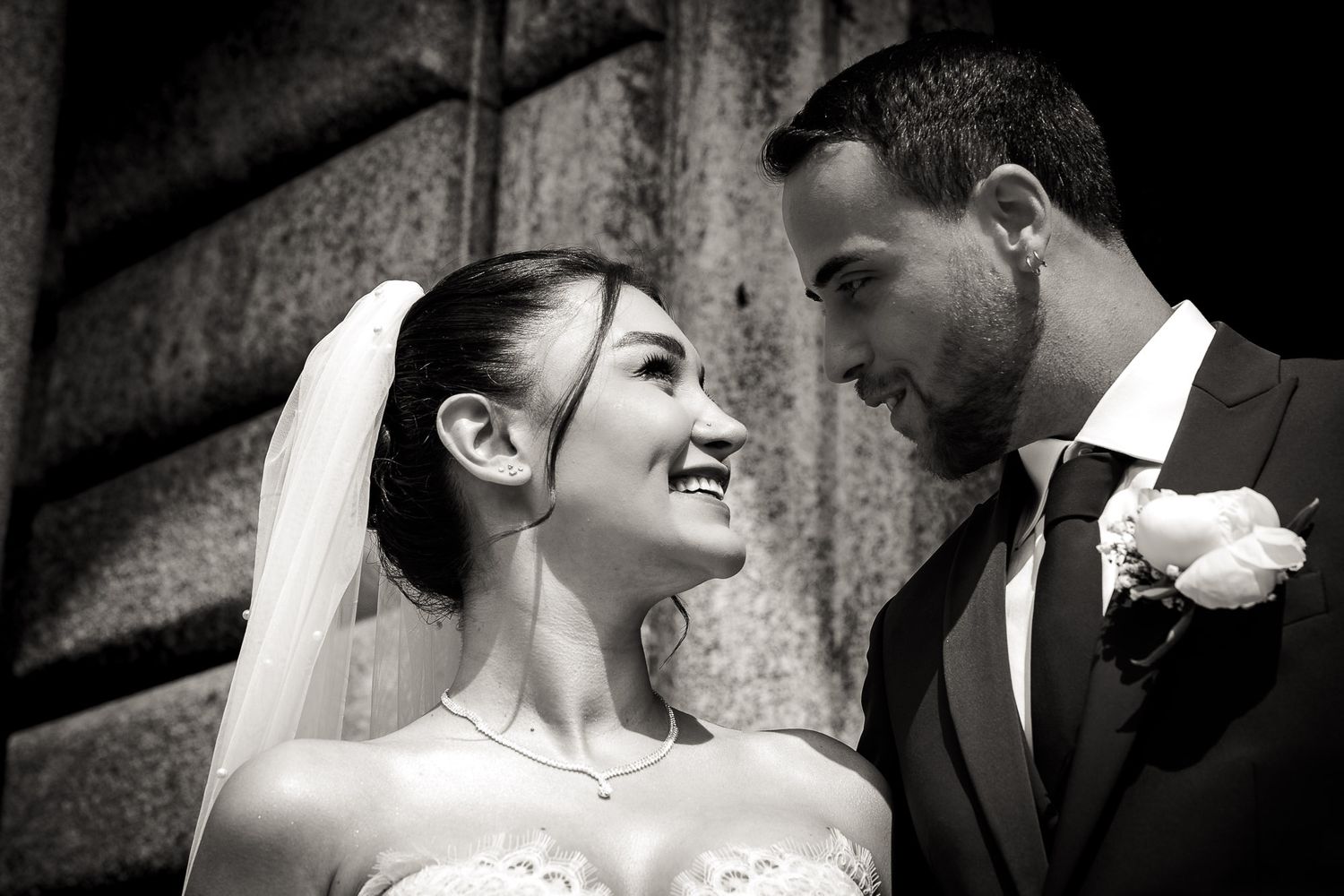 Black and white wedding photo of a smiling bride in veil and groom in suit gazing at each other outdoors.
