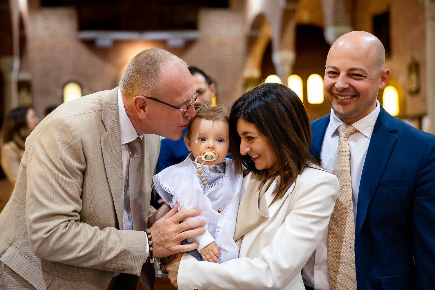 Family gathered in a church celebrating a baby baptism, with guests smiling and kissing the infant.