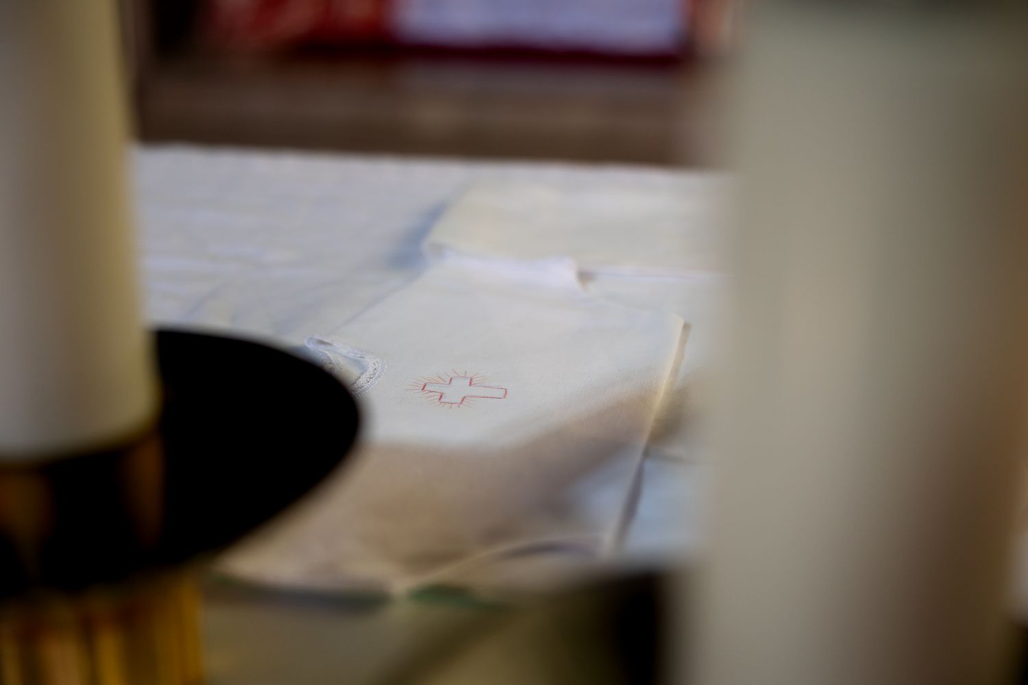 White linen cloth with red embroidered monogram on altar table with candles in soft focus background.