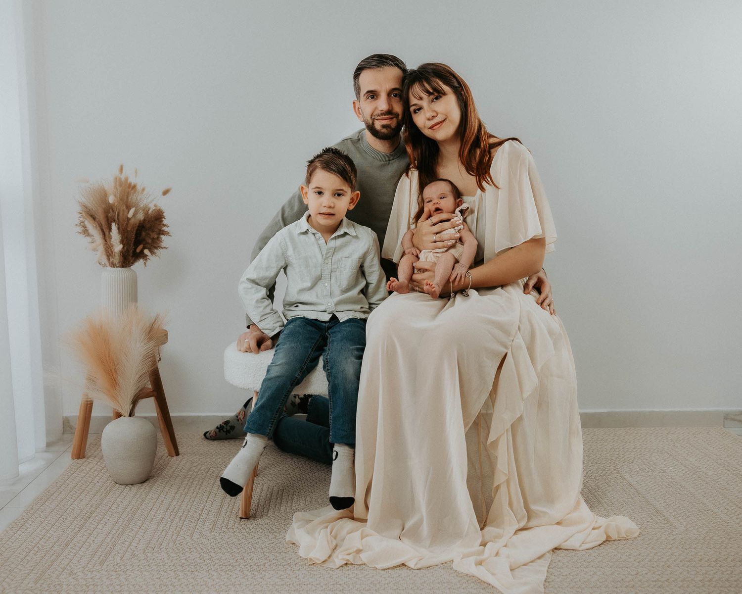 Family portrait with parents, toddler son, and newborn baby posing together in a bright, minimalist studio setting.