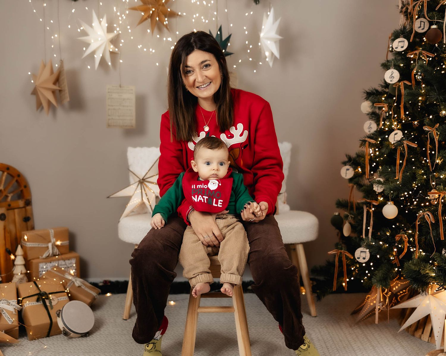 A mother and baby in matching red reindeer Christmas sweaters pose together near a decorated tree and wrapped gifts.