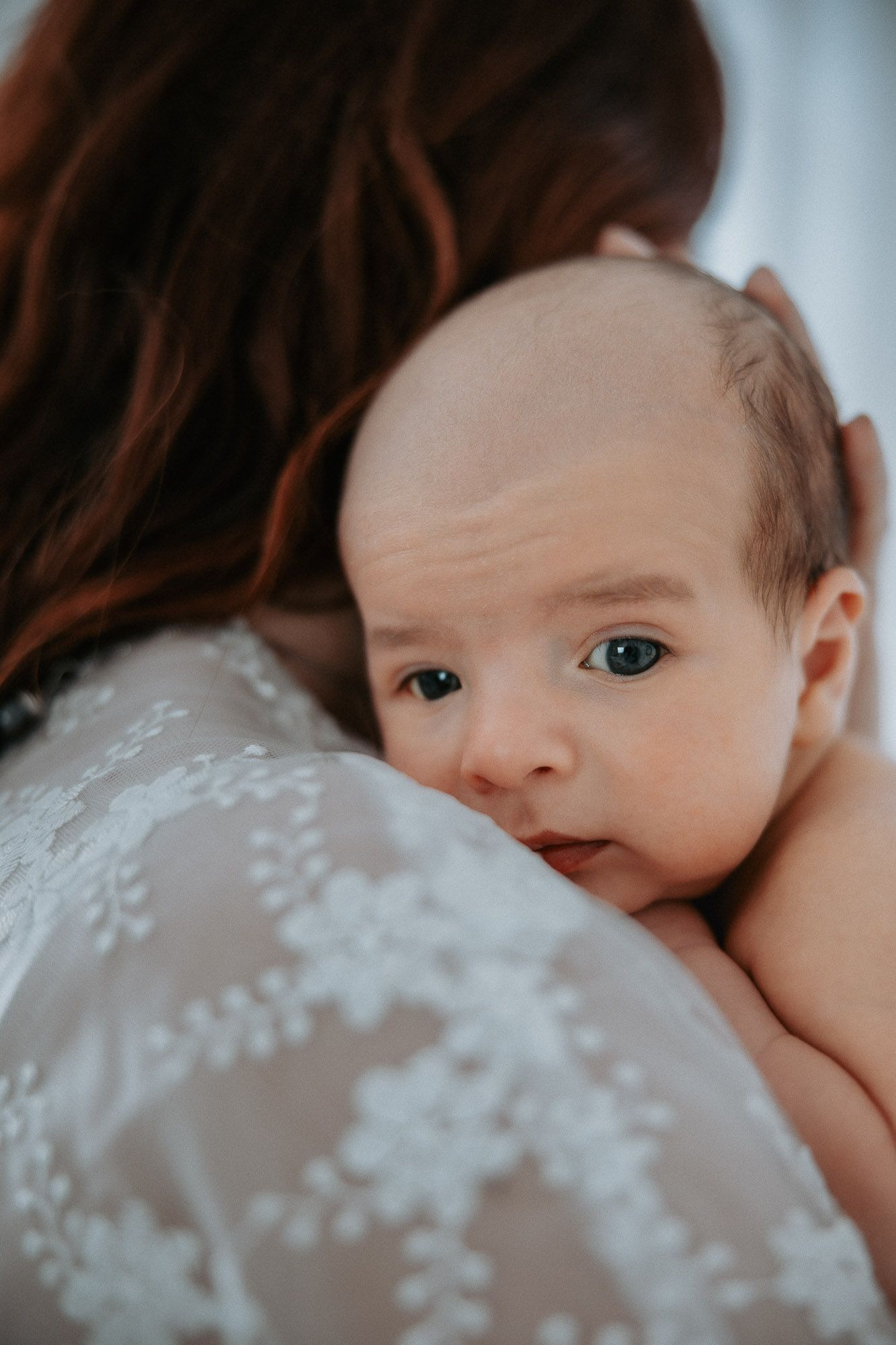 A newborn baby with blue eyes rests on a mother's shoulder, wearing a white lace dress, in a tender moment.