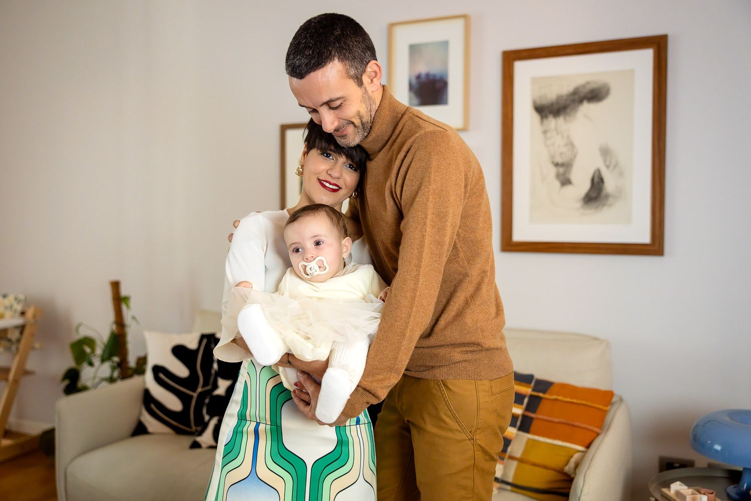 Father holding baby while young child hugs his legs in a warm, cozy home setting with framed art on walls.