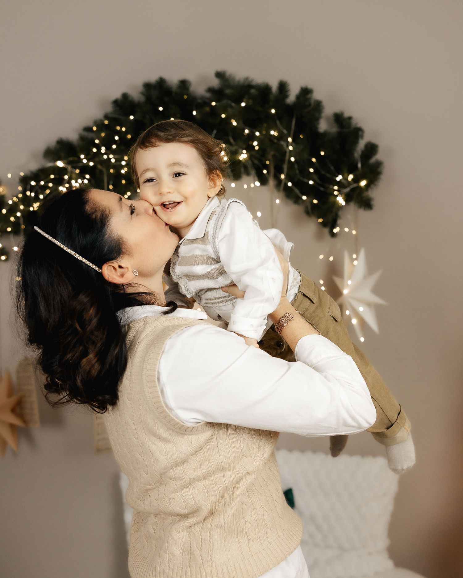 A mother kisses her smiling toddler while holding him up against a festive Christmas garland with warm fairy lights.