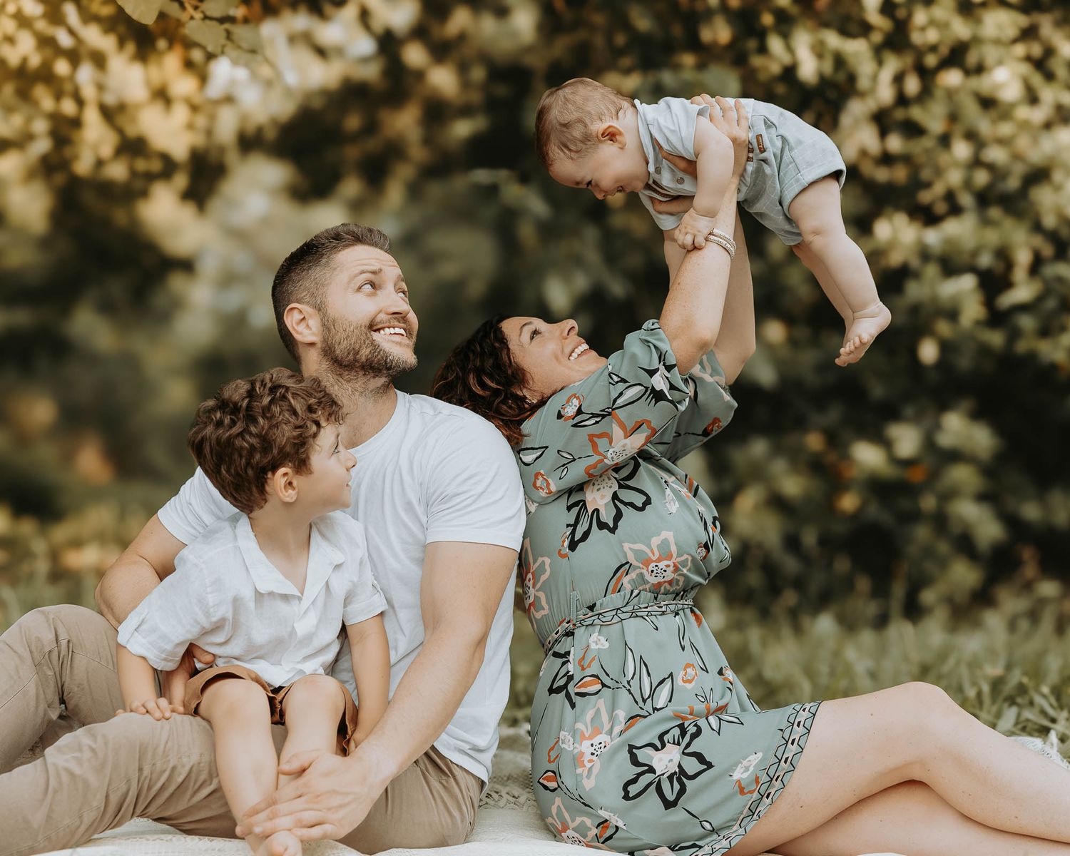 Happy family sitting outdoors in golden light, parents smiling as mother lifts baby while young son sits beside father.