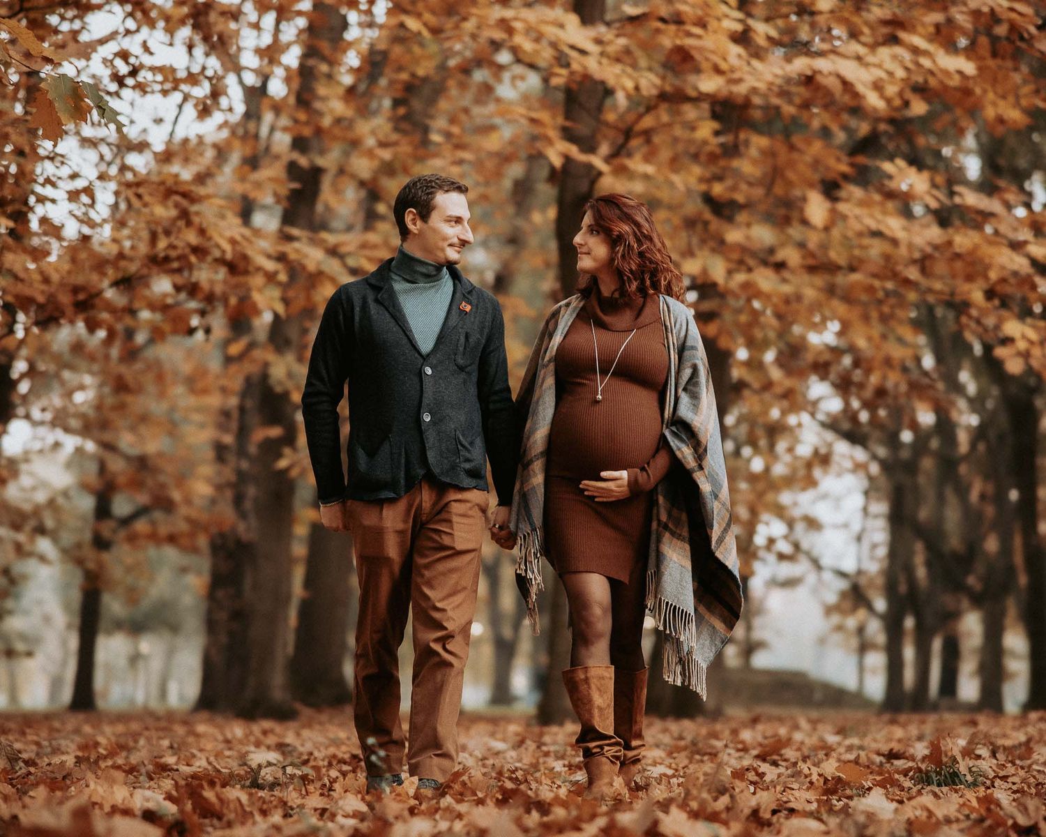 Couple walking in autumn park, pregnant woman in brown dress, man in navy blazer, surrounded by fall foliage.