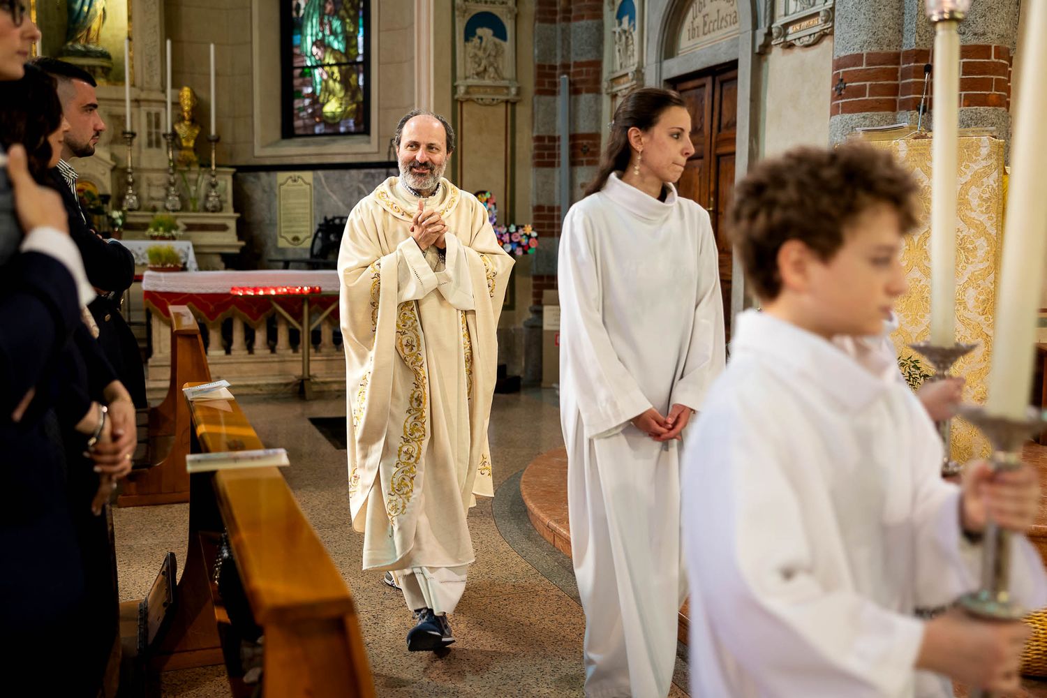 Catholic priest in white vestments leads altar servers in a church procession during a religious ceremony.