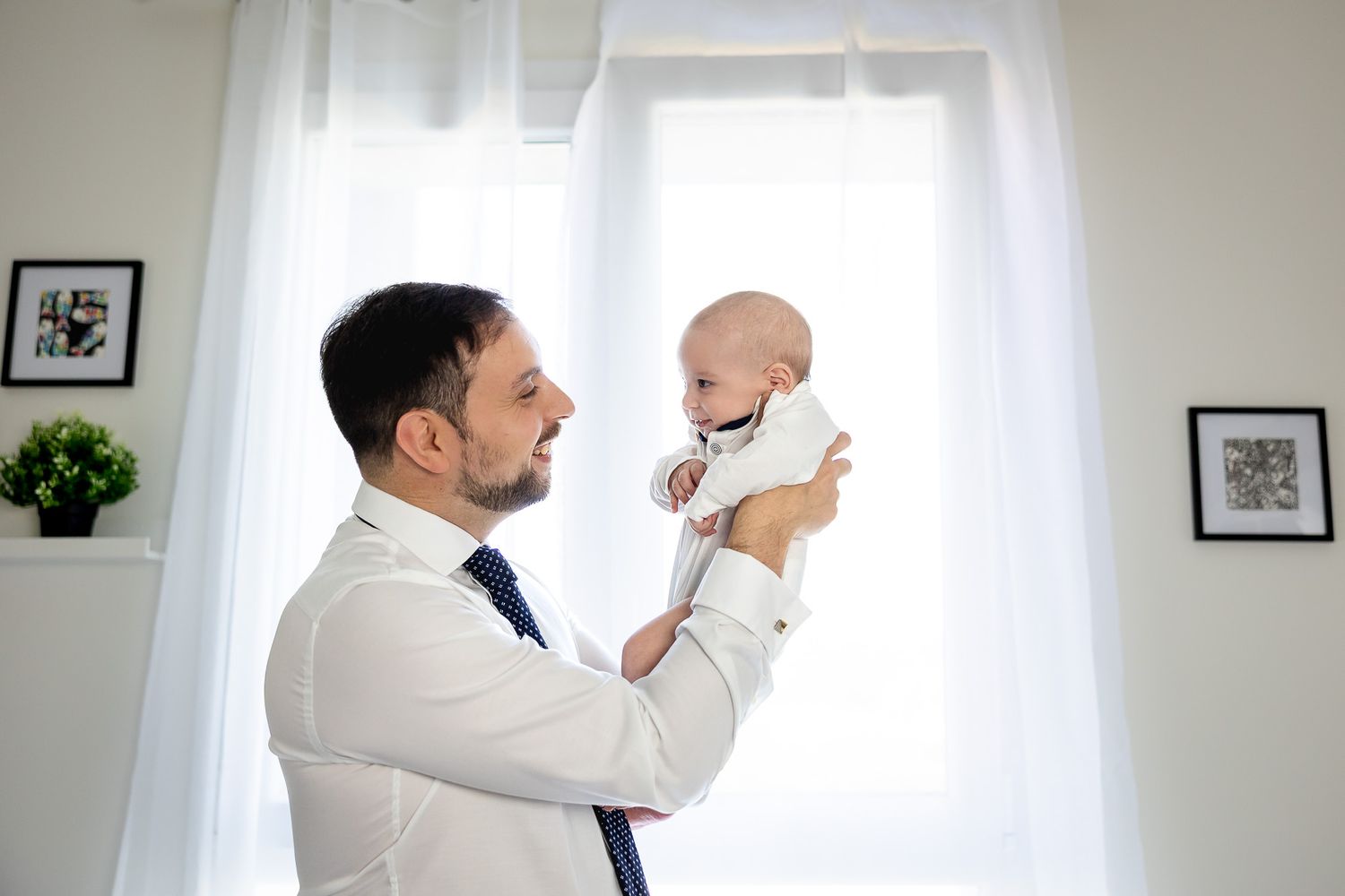 A well-dressed father in a white shirt and tie lovingly lifts his smiling baby in a bright, modern room.