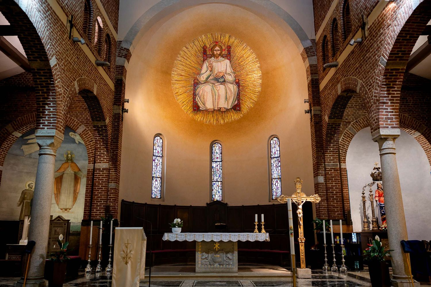 Catholic church interior with brick walls, stained glass windows, altar, and mosaic of Jesus Christ in golden halo above apse.