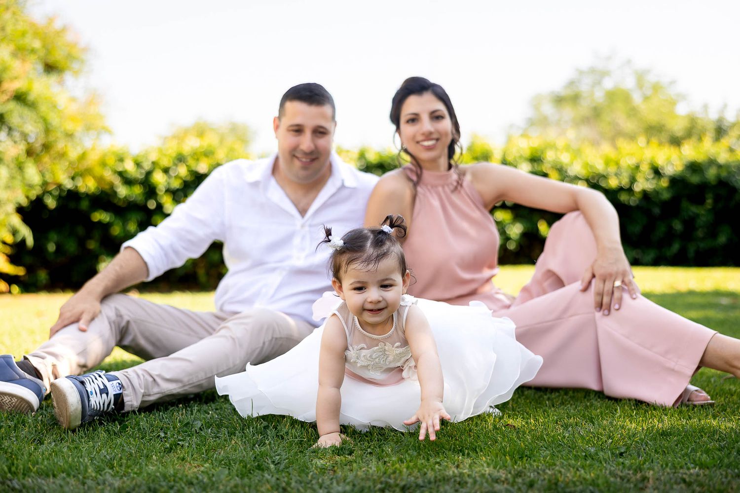 Happy family with parents sitting on green grass with smiling baby girl in white dress at outdoor photoshoot.